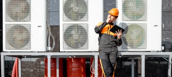Man holding tablet and wearing safety hat, stands in front of a rack of heat pumps