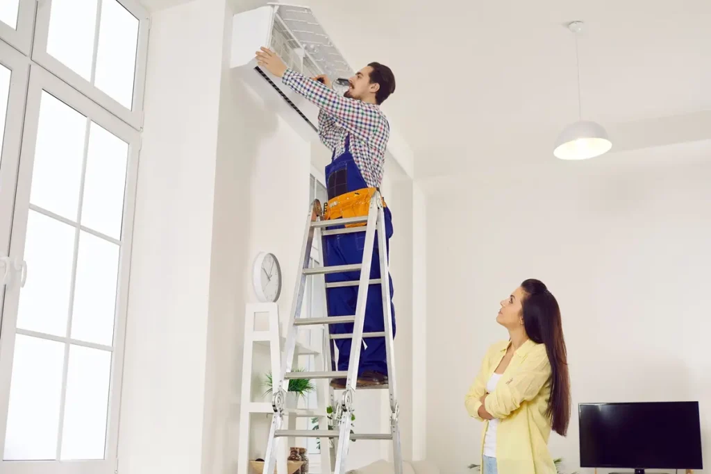Male technician on a ladder fixing an air conditioning unit while a woman stands nearby with folded arms.