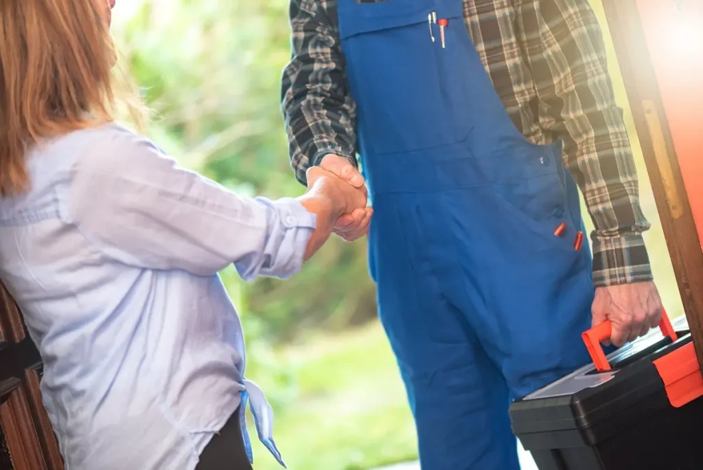 Repairman in blue overalls holding a toolbox in his left hand while shaking hands with his right with a a woman in a white shirt.