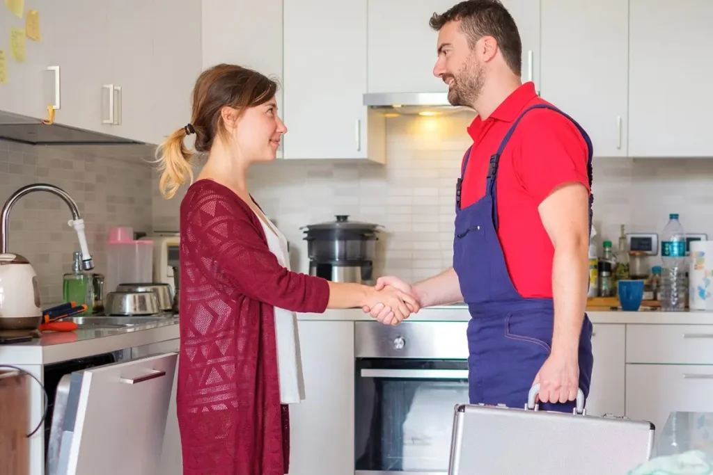 Technician in red shirt and denim work pants holding a briefcase in his left hand using his right to shake hands with a smiling woman in her kitchen