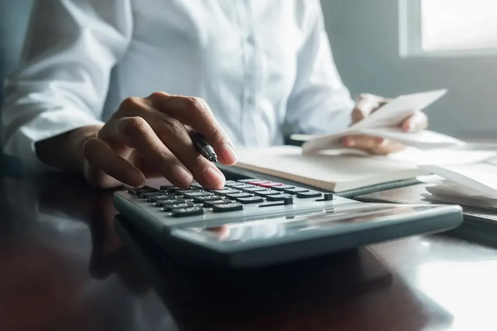 Woman in white dress shirt holding pen in one hand with her finger on a calculator and bills in another hand on a table with more bills scattered.