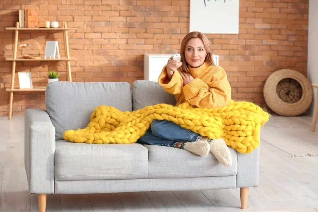 Smiling woman in yellow sweater sitting on sofa with yellow comforter holing a remote in hand with a decorative background of brick and wooden rack.